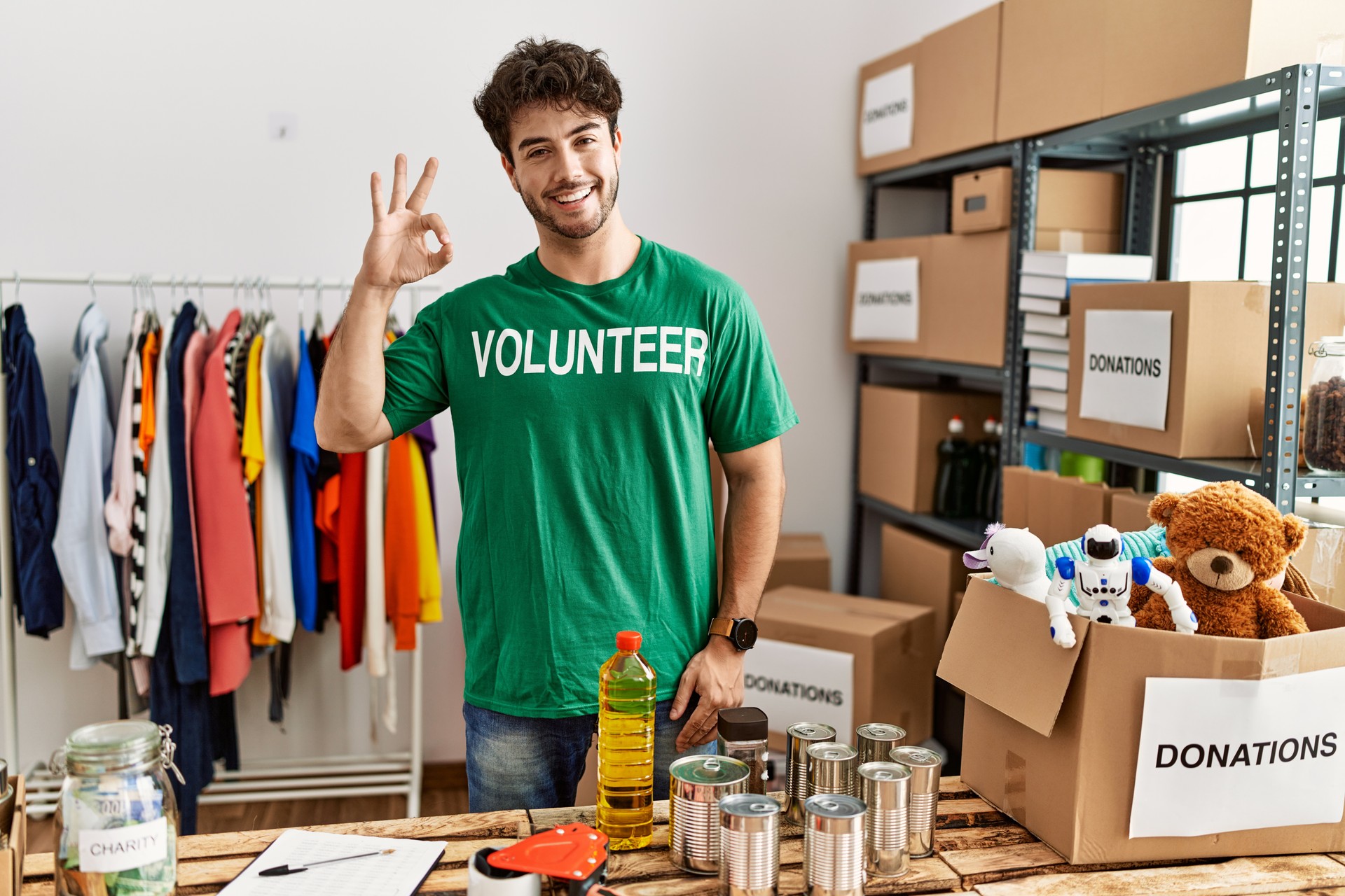 Hispanic man wearing volunteer t shirt at donations stand doing ok sign with fingers, smiling friendly gesturing excellent symbol